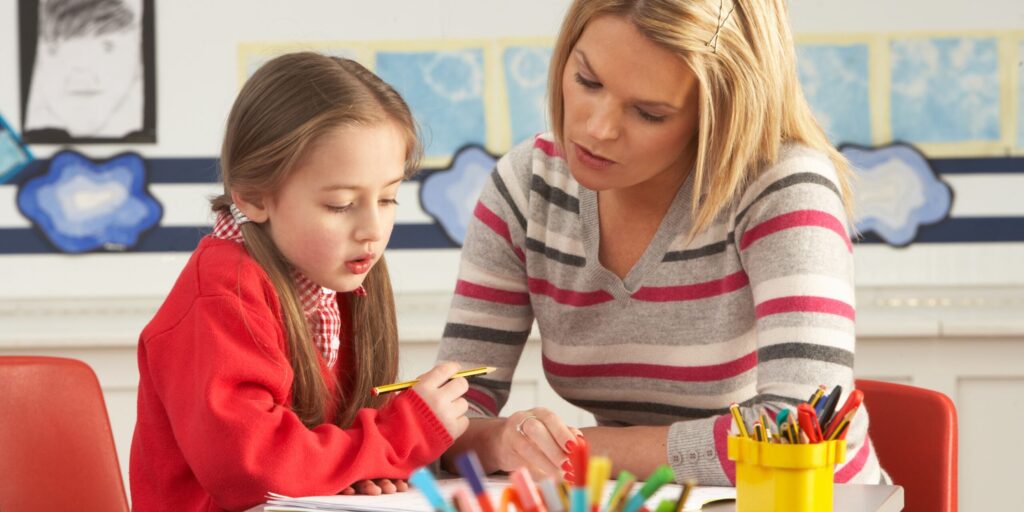 Female Primary School Pupil And Teacher Working At Desk In Classroom